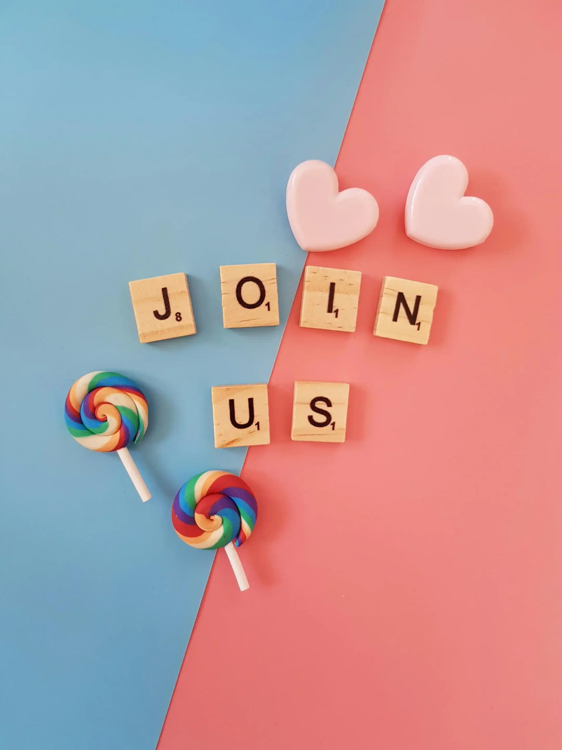 Colorful flat lay with Scrabble tiles spelling “JOIN US,” two heart shapes and two rainbow lollipops on a split blue and pink background, conveying a friendly invitation—ideal for a vibrant “Contact Us” or community engagement page.