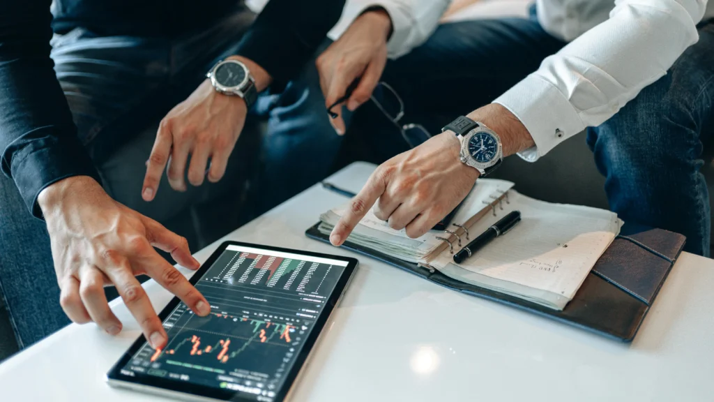 Close-up of business professionals pointing at a tablet displaying cryptocurrency trading charts, with notebooks and pens on the table, suggesting a strategy meeting.
