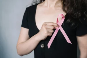 Woman in a black dress holding a pink awareness ribbon near her chest, symbolizing support for cancer-related causes, such as mesothelioma awareness.