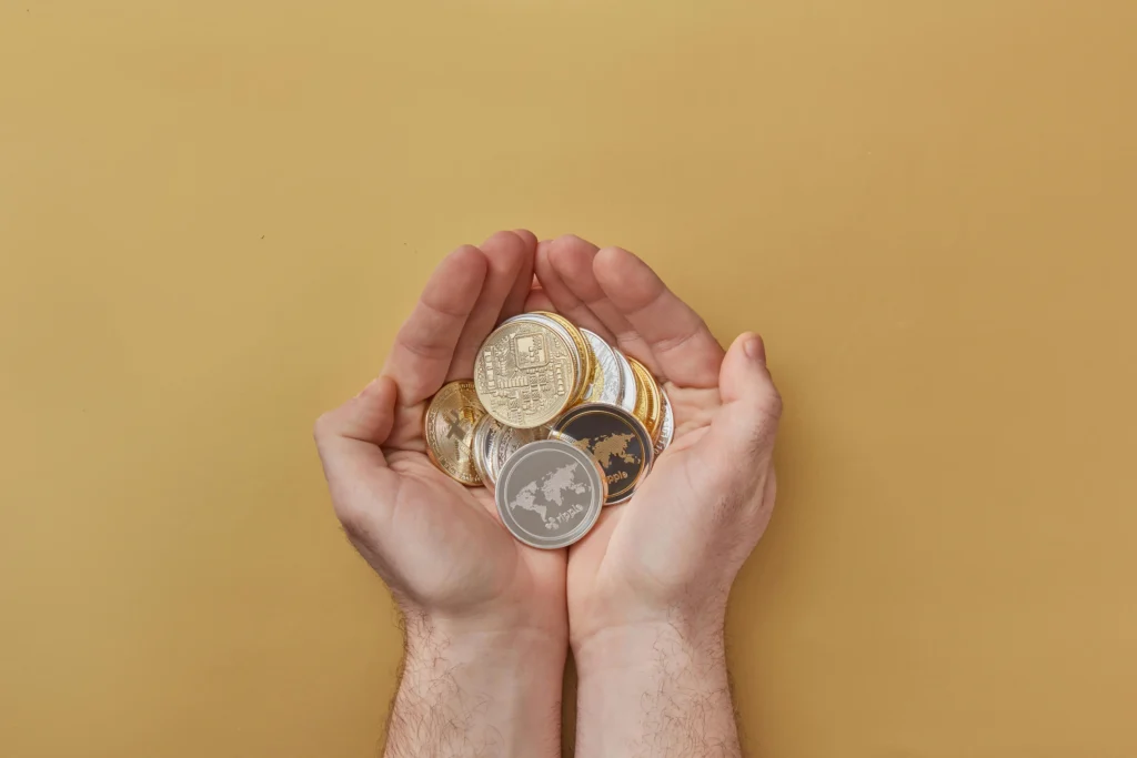 A pair of hands holding a variety of physical cryptocurrency coins, including Bitcoin and Ripple, over a neutral beige background.