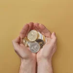 A pair of hands holding a variety of physical cryptocurrency coins, including Bitcoin and Ripple, over a neutral beige background.