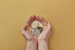A pair of hands holding a variety of physical cryptocurrency coins, including Bitcoin and Ripple, over a neutral beige background.