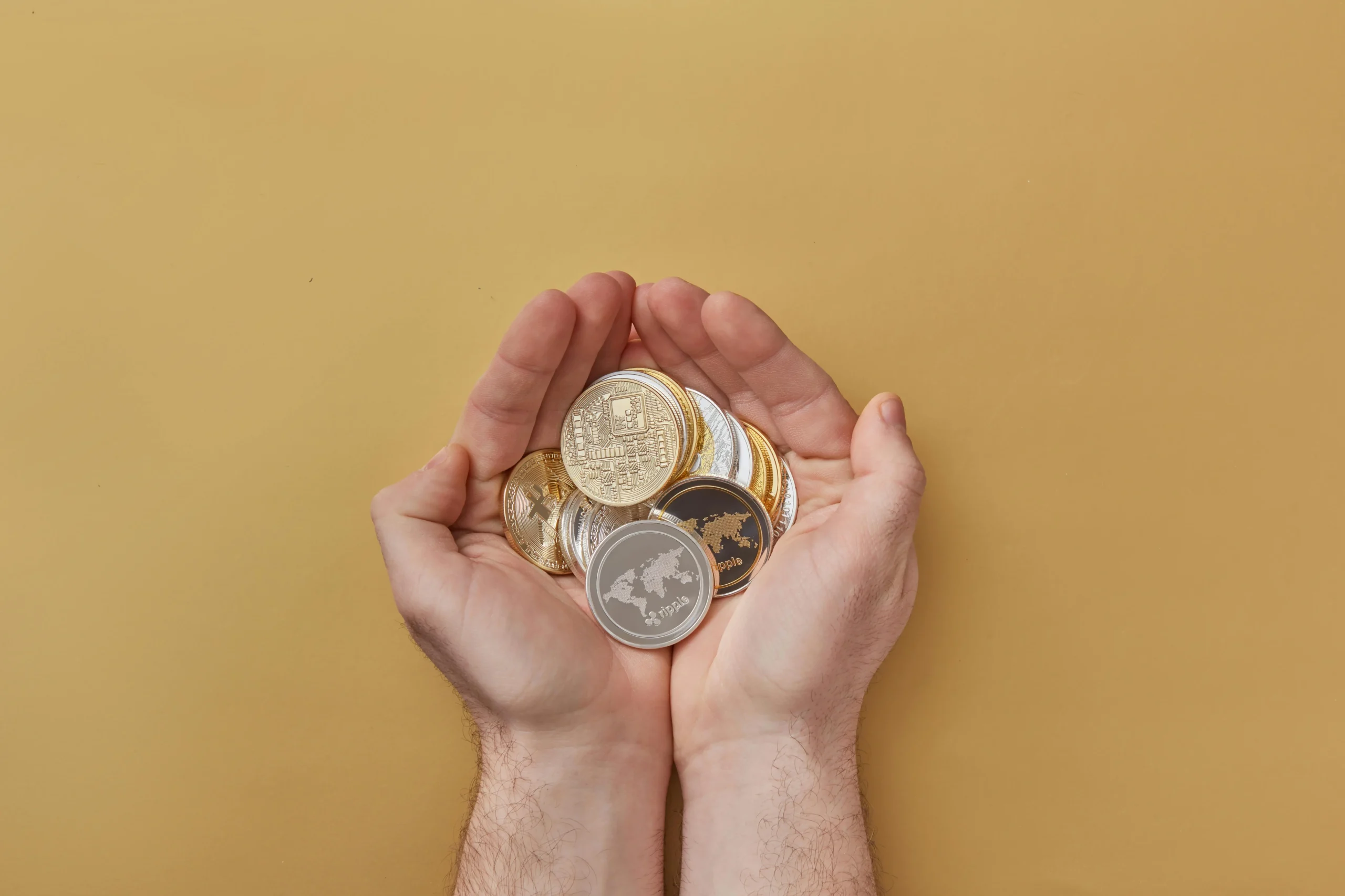 A pair of hands holding a variety of physical cryptocurrency coins, including Bitcoin and Ripple, over a neutral beige background.