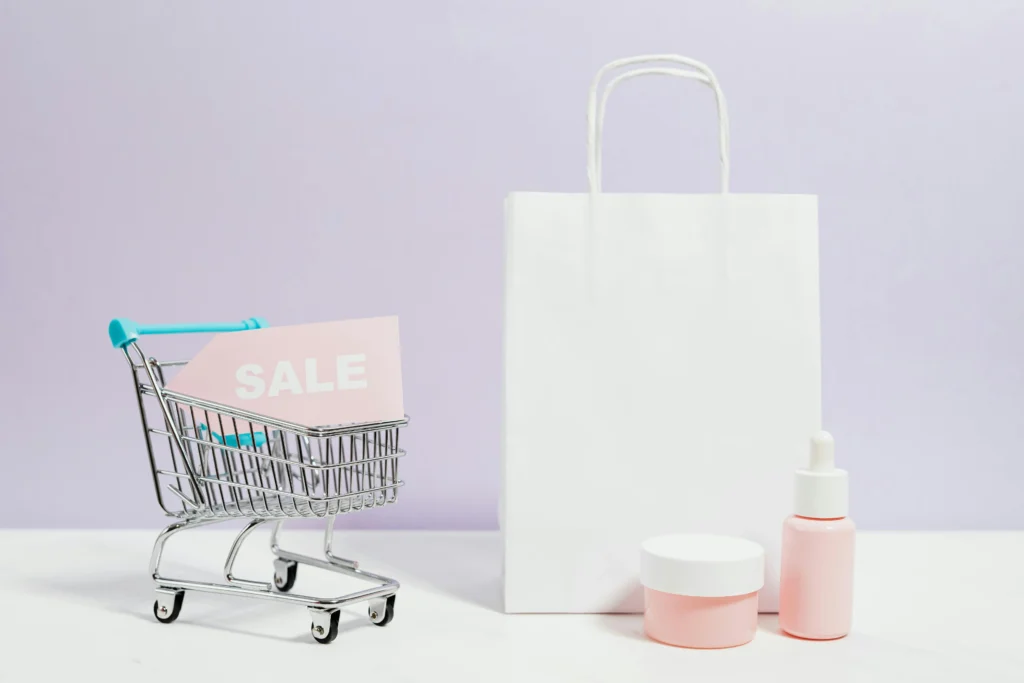 A small shopping cart holding a pink “SALE” sign beside beauty products and a white paper bag, representing branding and promotion in ecommerce.