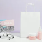 A small shopping cart holding a pink “SALE” sign beside beauty products and a white paper bag, representing branding and promotion in ecommerce.
