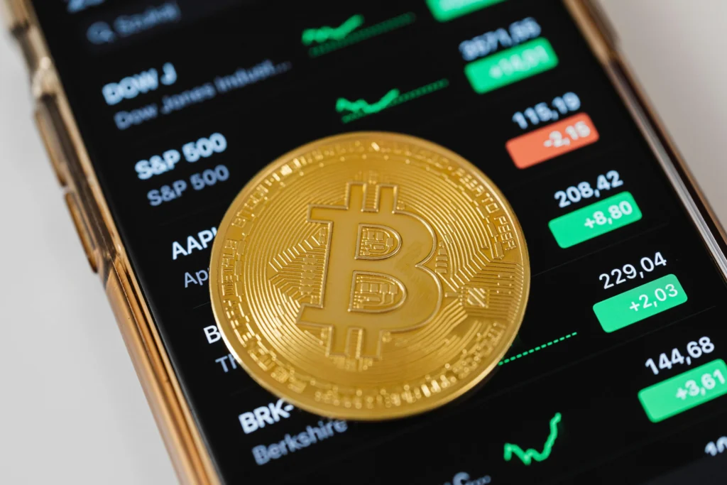 "A golden Bitcoin coin placed on a smartphone screen displaying stock market indexes and price changes, indicating financial market activity."