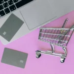 Online shopping concept with laptop, credit cards, and miniature shopping cart on a pink background — representing the evolution of e-commerce.