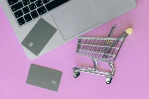 Online shopping concept with laptop, credit cards, and miniature shopping cart on a pink background — representing the evolution of e-commerce.