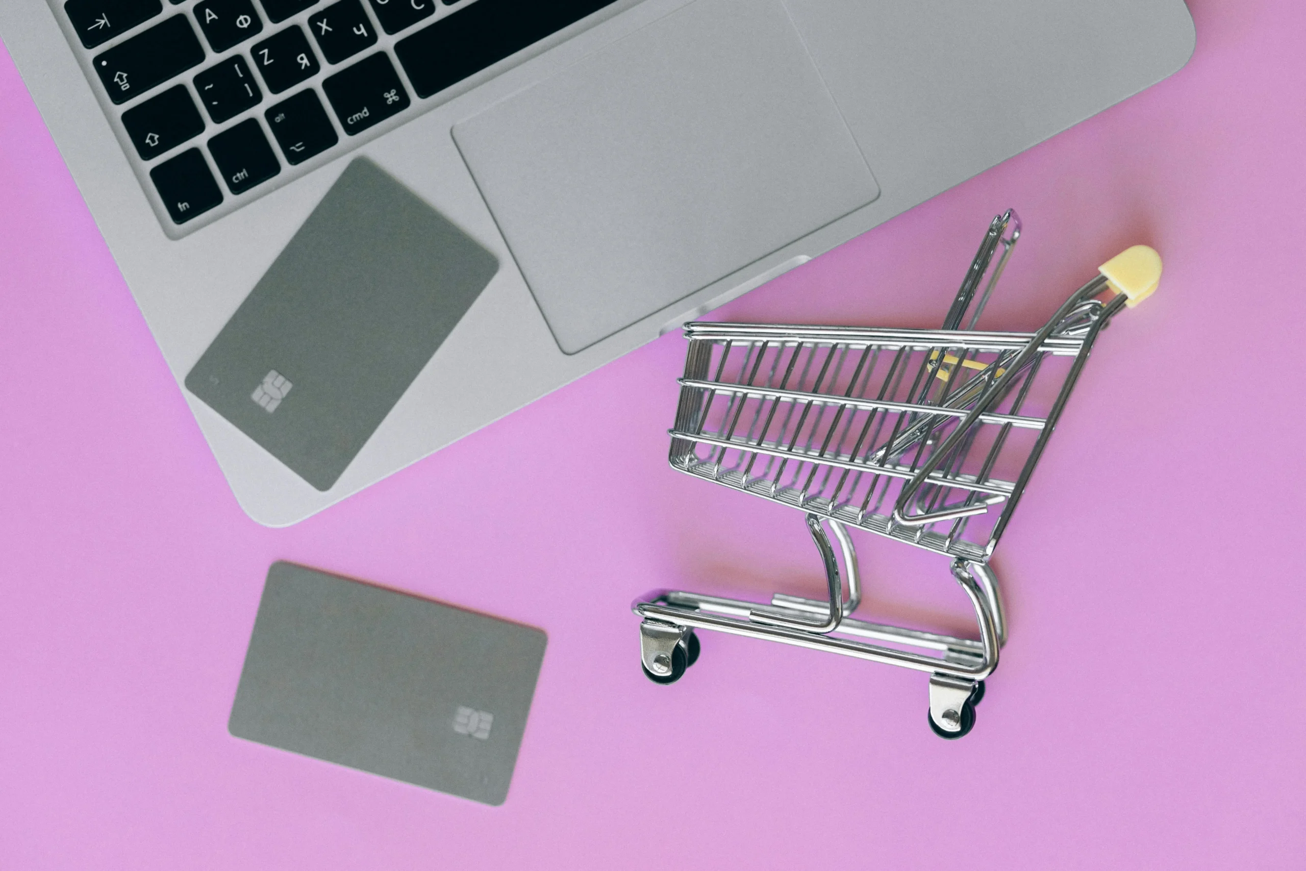 Online shopping concept with laptop, credit cards, and miniature shopping cart on a pink background — representing the evolution of e-commerce.