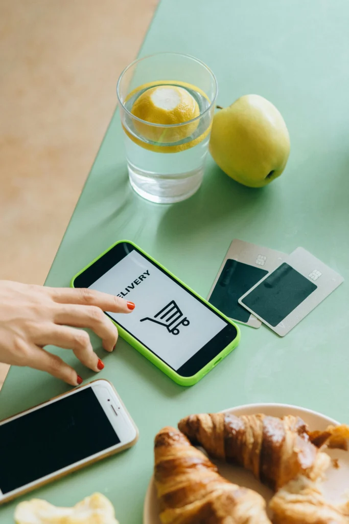 Person shopping online using a smartphone app at a breakfast table, with credit cards and food nearby—depicting convenient mobile commerce.